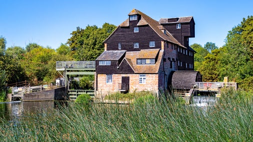 An historic riverside watermill, built of red brick and black boards. It is summer in the photo, the sky is blue and the riverbank is green with vegetation